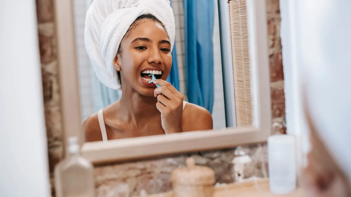 Reflection of young African American female with white towel on head looking in mirror while cleaning teeth with dental flosser